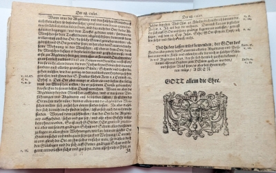 Atvērta vēsturiska grāmata gotiskajā šriftā, kuras labajā lappusē redzams dekoratīvs baroka stila grafikas elements un uzraksts "GOTT allein die Ehre".