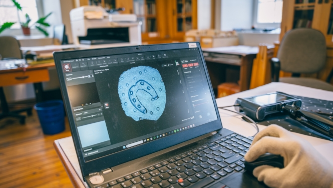 Roka baltā cimdā vada datorpeli, ekrānā redzams arheoloģiska priekšmeta (pakava formas saktas) 3D skenēšanas process speciālā programmā.