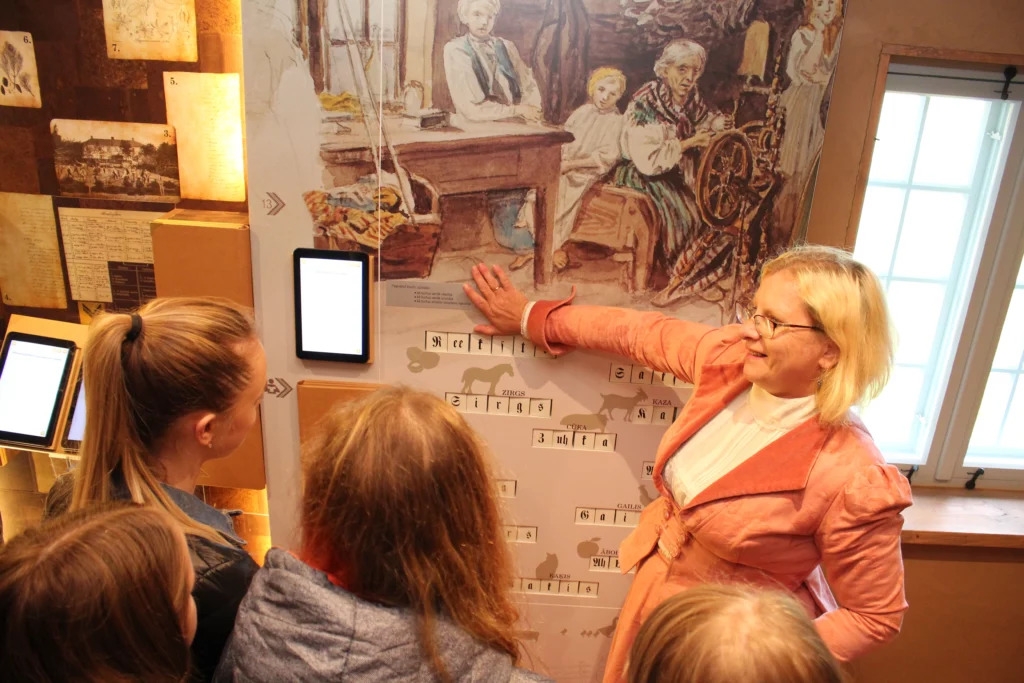 Izglītojošā programma Latviešu valoda – manas valsts valoda. Vadītāja rāda skolēnu grupai senu attēlu latviešu ģimeni. 