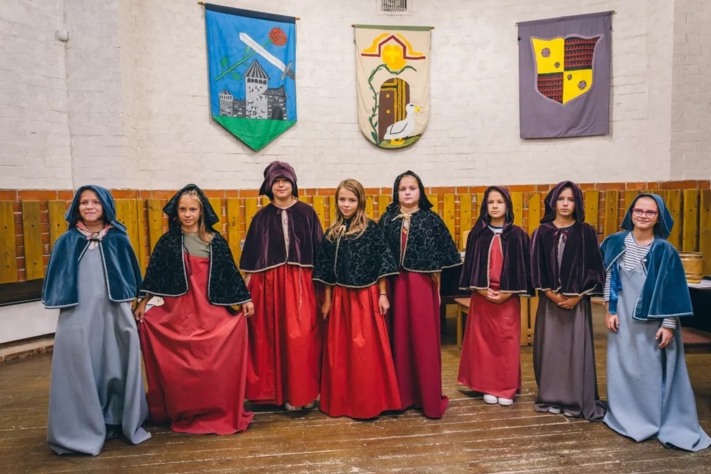 Ciemos pie Turaidas fogta. Skolnieču grupa stāv kājās spilgtos viduslaiku tērpos