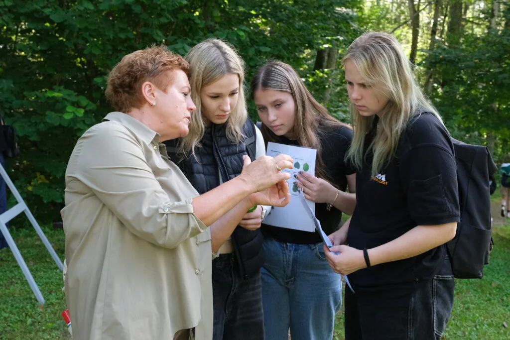 Lapkoku daudzveidības salīdzināšana Turaidas muzejrezervāta meža parkā un Lībiešu takā 7.–9. kl. skolēniem. Vadītāja rāda trīs meitenēm koka lapu.
