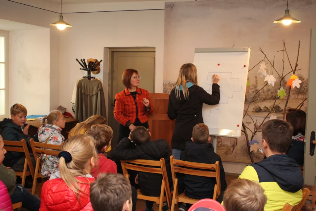 Muzejpedagoģiskās programma Uzvārdu došana Vidzemē. Vadītāja un skolnieku grupa skatās, kā skolniece raksta uz tāfeles.