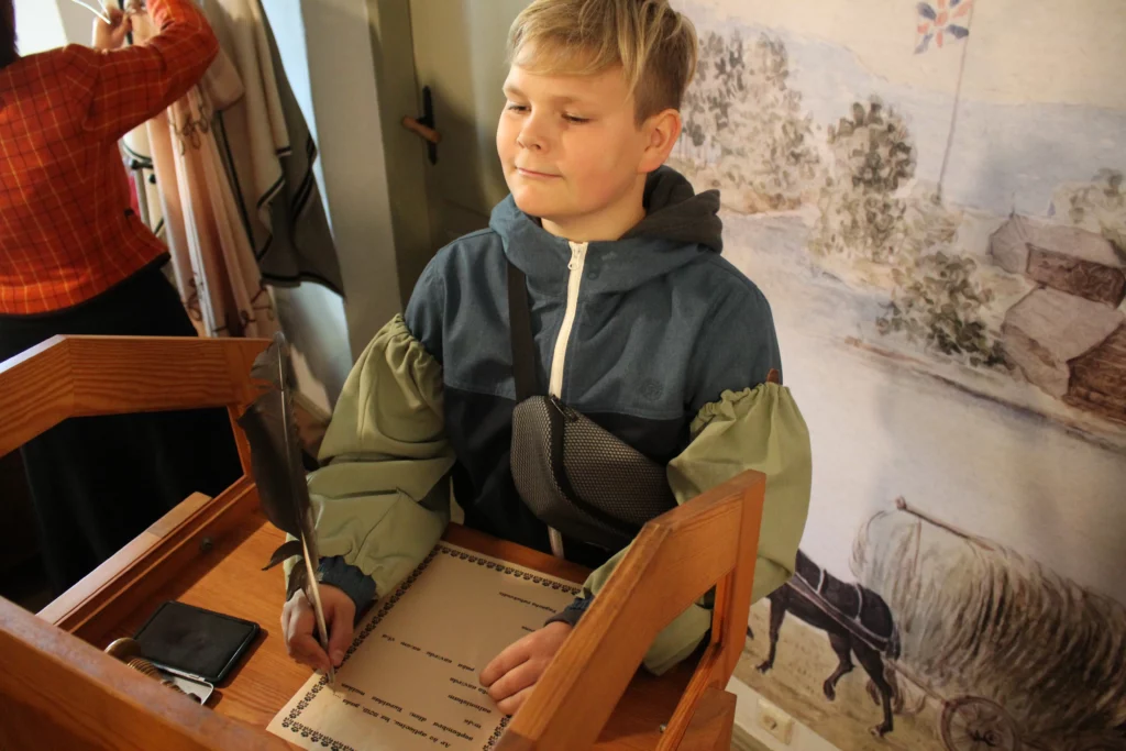 Muzejpedagoģiskās programma Uzvārdu došana Vidzemē Skolnieks raksta ar tinti un putna spalvu uz baltas lapas.