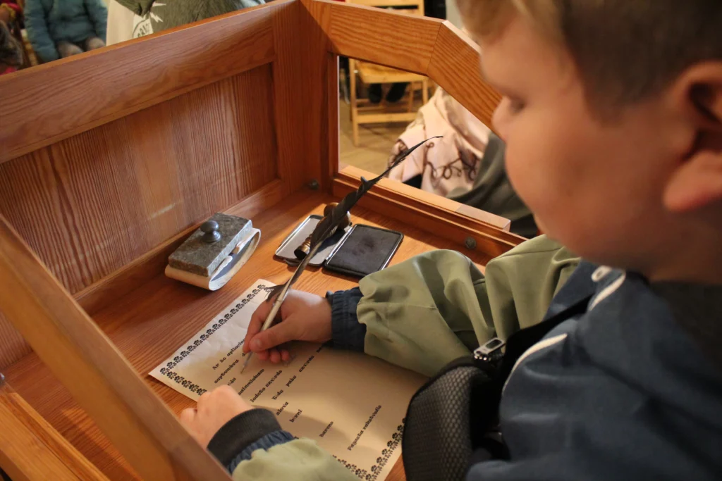 Muzejpedagoģiskās programmas Uzvārdu došana Vidzemē Skolnieks raksta uz lapas ar tinti un putna spalvu