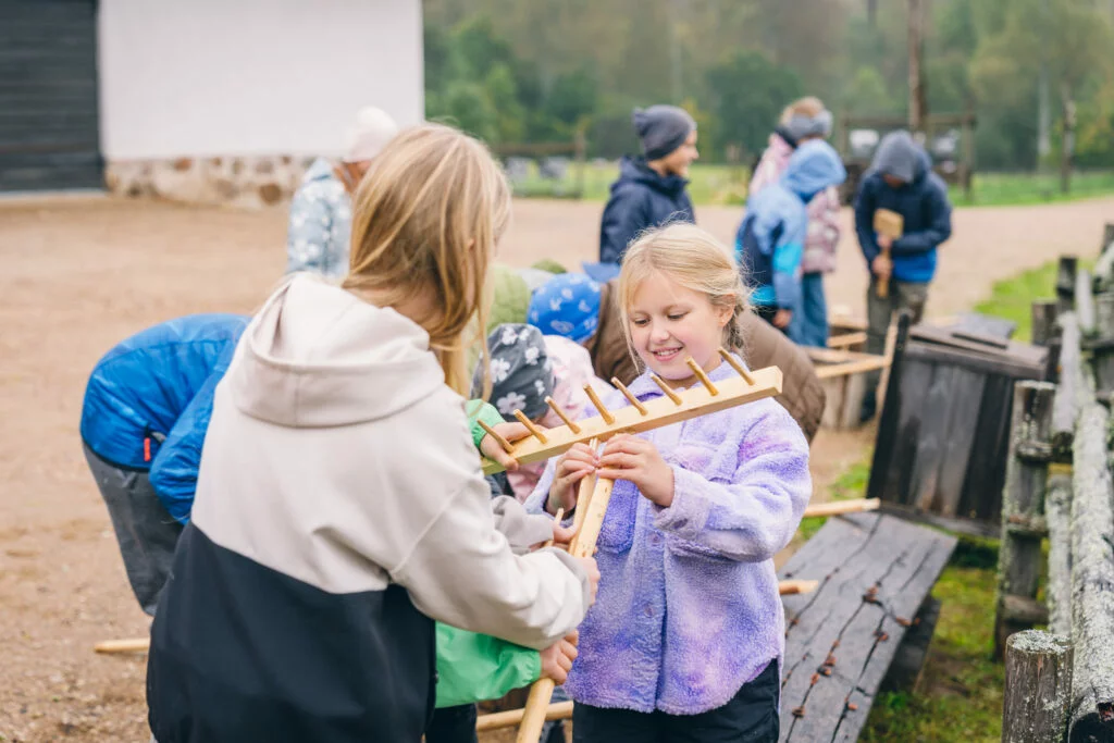 Bērni izglītojošā programmā Kas ir klaušinieks Divas meitenes apskata koka grābekli.