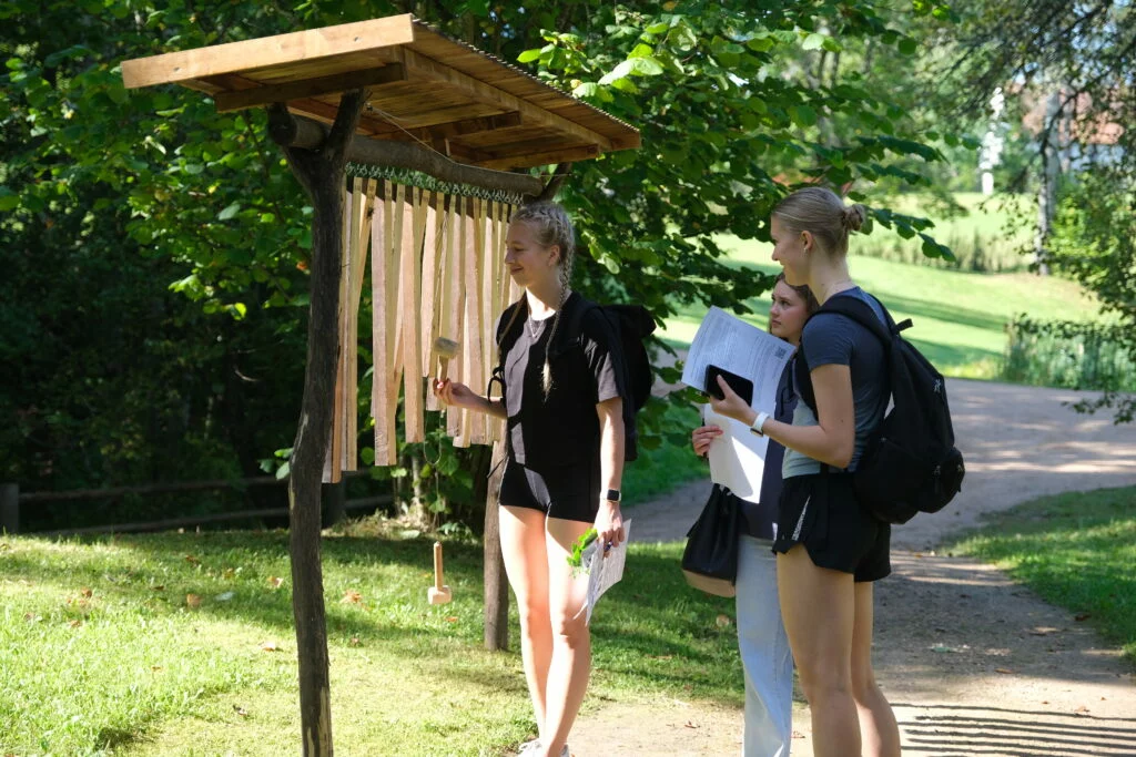 Koksnes krājas salīdzināšana kokiem Turaidas muzejrezervāta meža parkā 7.–9. klašu skolēniem. Trīs meitenes pēta koka zarus.