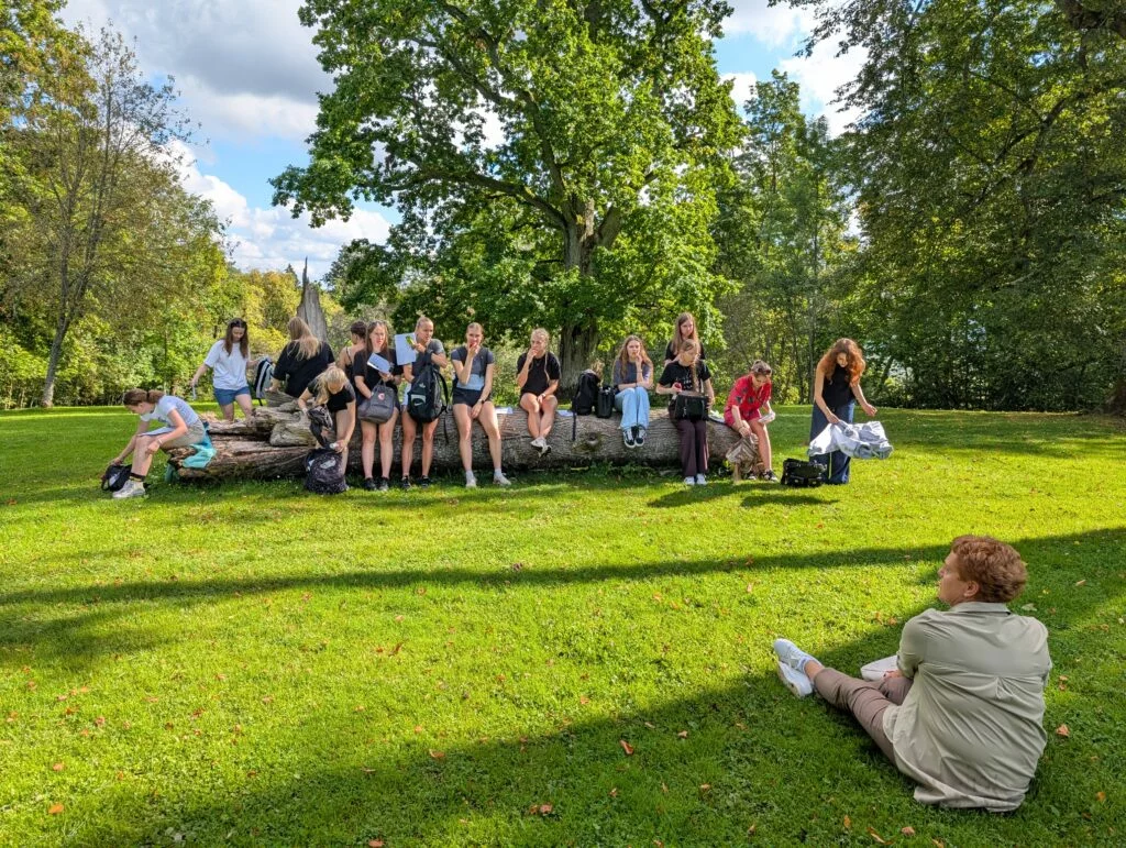 Koksnes krājas salīdzināšana kokiem Turaidas muzejrezervāta meža parkā (10.–12. kl.). Jauniešu grupa sēž Turaidas muzejrezervāta meža parkā un klausās vadītājas stāstīto.