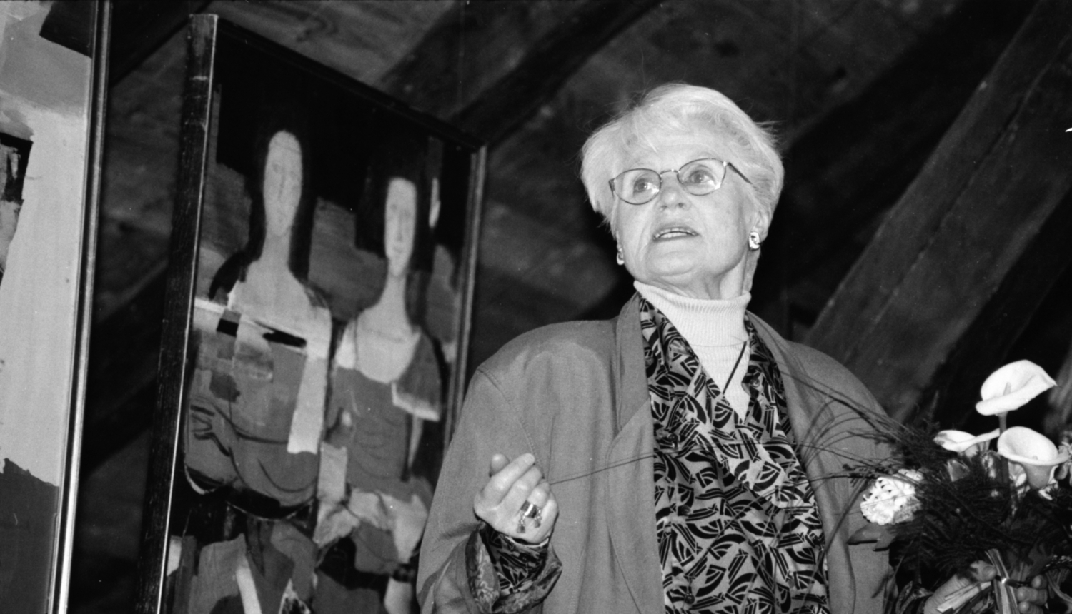 Melnbalta fotogrāfija, kurā māksliniece Džemma Skulme uzstājas; fonā redzama viena no viņas raksturīgajām gleznām ar stilizētām sieviešu figūrām.