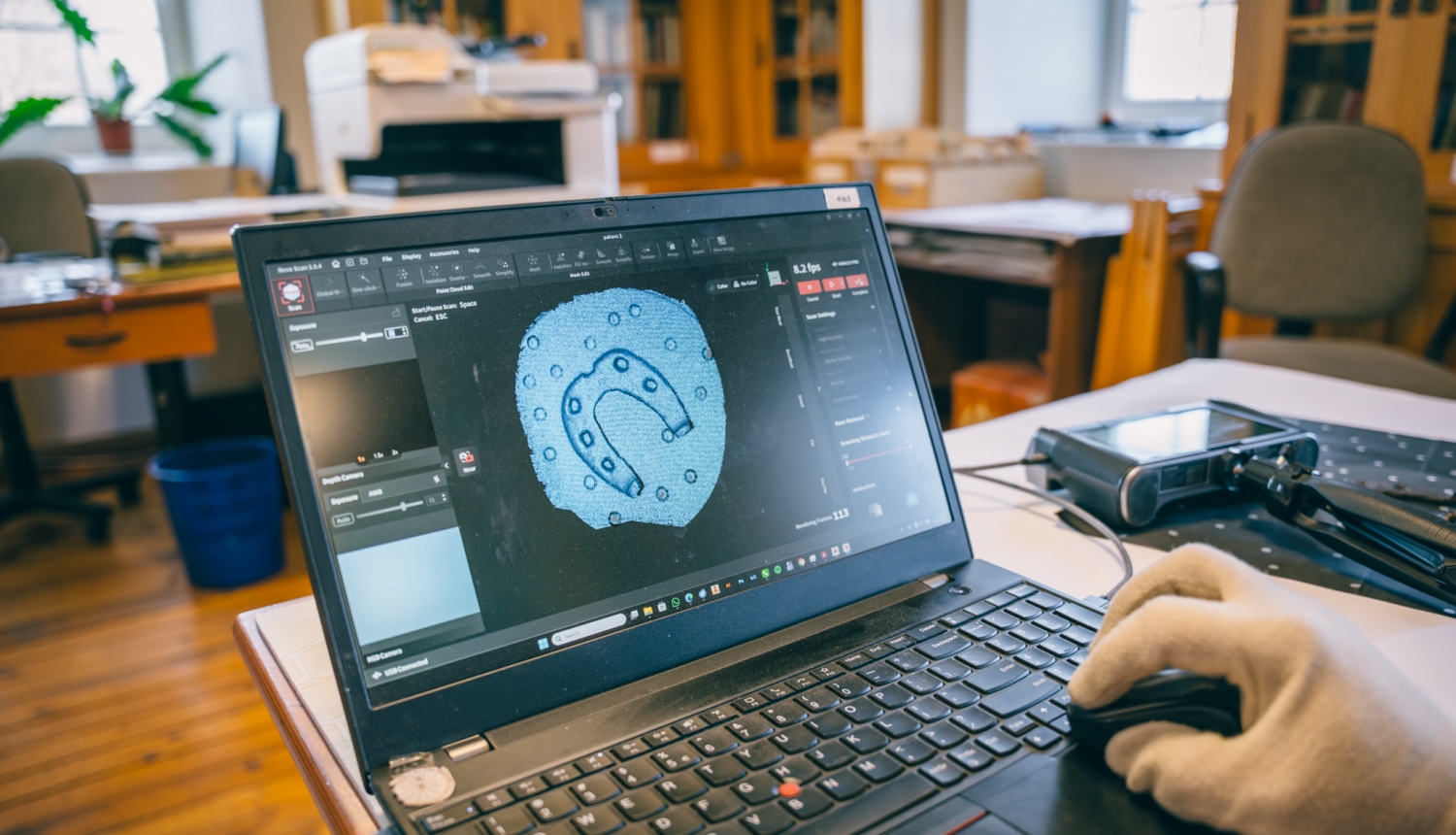 Roka baltā cimdā vada datorpeli, ekrānā redzams arheoloģiska priekšmeta (pakava formas saktas) 3D skenēšanas process speciālā programmā.