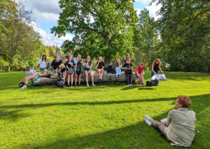 Koksnes krājas salīdzināšana kokiem Turaidas muzejrezervāta meža parkā (10.–12. kl.). Jauniešu grupa sēž Turaidas muzejrezervāta meža parkā un klausās vadītājas stāstīto.